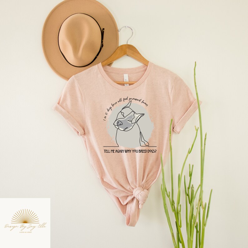 May include: A light pink t-shirt with a dog graphic and the text "I'm 10 dogs have all found permanent homes. Tell me again why you breed dogs?". A tan hat and a wooden hanger are behind the shirt.