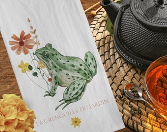 Puede incluir: Paño de cocina blanco con una ilustración de acuarela de una rana verde y flores marrones y amarillas. El texto "LA GRENOUILLE DU JARDIN" está impreso en marrón. Una tetera negra y una taza de té están en el fondo.