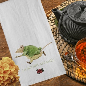 May include: White cotton kitchen towel featuring a watercolor illustration of a running mouse in a green jacket and tan pants. The text "LA SOURIS AFFAMÉE" is printed in green. A sprig of red berries is below the text. A yellow flower and a black teapot are in the background.