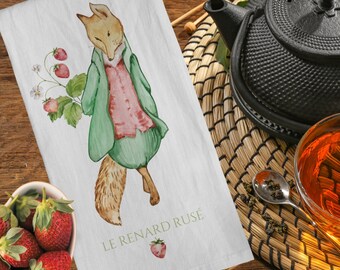 Op de afbeelding: Witte keukendoek met een aquarel illustratie van een vos in een groene jas en roze shirt, met aardbeien. De tekst "LE RENARD RUSÉ" is eronder gedrukt. Een theepot en aardbeien zijn ook zichtbaar.