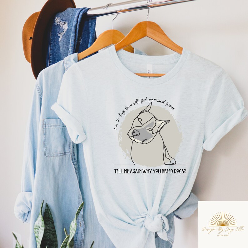 May include: Light blue t-shirt with a line drawing of a dog and the text "I in 10 dogs born will find permanent homes. TELL ME AGAIN WHY YOU BREED DOGS?" The shirt is hanging on a wooden hanger.