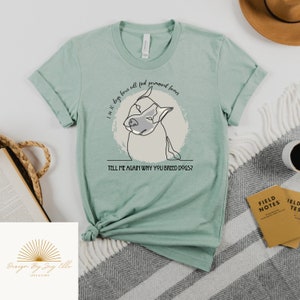 May include: A light green t-shirt with rolled-up sleeves. The shirt features a line drawing of a dog's face inside a light gray circle. Text on the shirt reads, "I am 10 dogs born will find permanent homes. Tell me again why you breed dogs?"
