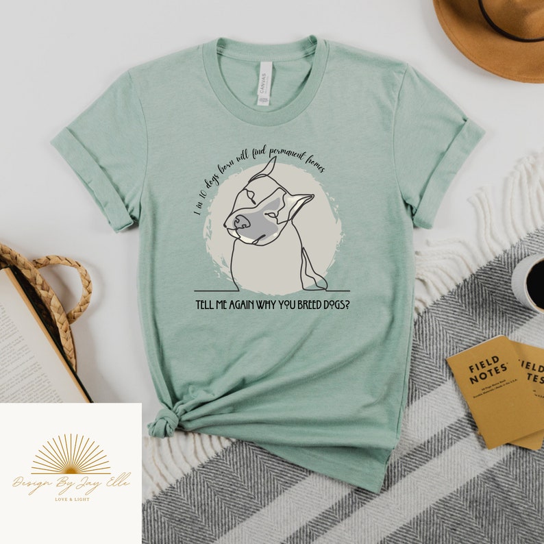 May include: A light green t-shirt with rolled-up sleeves. The shirt features a line drawing of a dog's face inside a light gray circle. Text on the shirt reads, "I am 10 dogs born will find permanent homes. Tell me again why you breed dogs?"