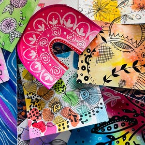 May include: A collection of colourful, hand-painted cards featuring floral and abstract designs. The cards are arranged in a pile, with a pink and white arch-shaped card on top.
