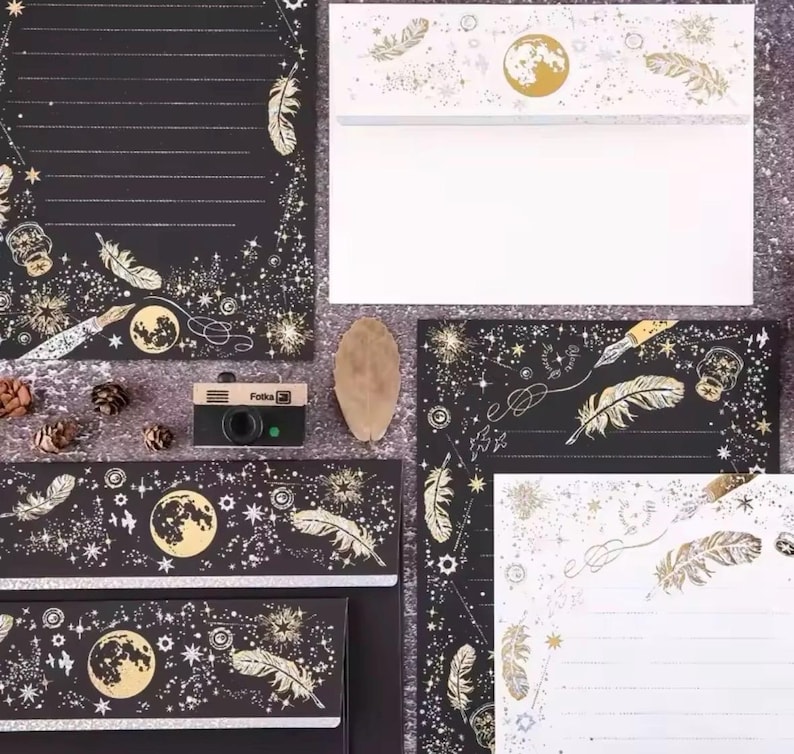 Puede incluir: Juego de papeler&iacute;a con dise&ntilde;o celestial. Fondo negro con detalles dorados de plumas, lunas y estrellas. Incluye sobres, papel de carta y una peque&ntilde;a c&aacute;mara.