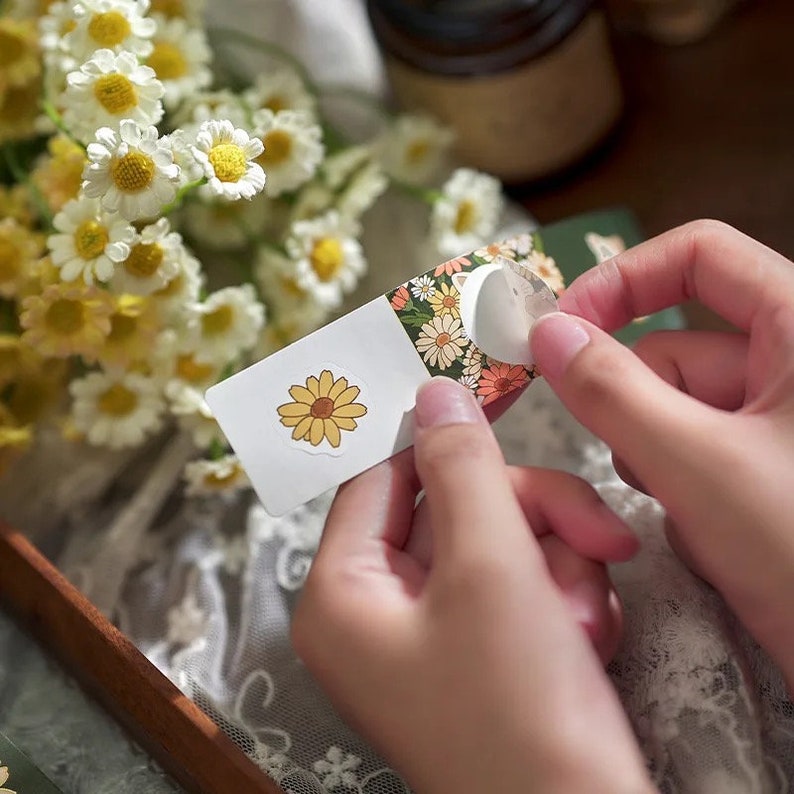 Pu&ograve; includere: Un foglio di adesivi con un design floreale. Gli adesivi presentano margherite gialle e arancioni su uno sfondo bianco. Il foglio viene staccato per rivelare un adesivo con un'unica margherita.