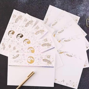 Puede incluir: Un juego de papeler&iacute;a con papel y sobres blancos, decorados con dise&ntilde;os celestiales dorados. Los dise&ntilde;os incluyen lunas, plumas y estrellas. Un bol&iacute;grafo dorado descansa en la esquina inferior izquierda.