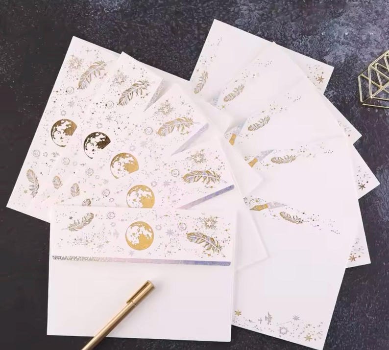 Puede incluir: Un juego de papeler&iacute;a con papel y sobres blancos, decorados con dise&ntilde;os celestiales dorados. Los dise&ntilde;os incluyen lunas, plumas y estrellas. Un bol&iacute;grafo dorado descansa en la esquina inferior izquierda.
