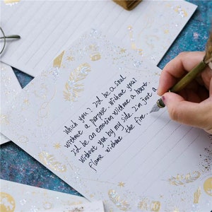 Puede incluir: Papel de escribir blanco con dise&ntilde;os de plumas y estrellas doradas. El papel tiene l&iacute;neas para escribir y contiene texto manuscrito. Una mano sostiene un bol&iacute;grafo, escribiendo en el papel. El texto dice: "Which you? I'd be a soul without a purpose..."