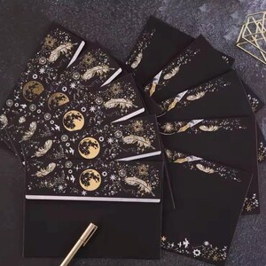 Puede incluir: Juego de papeler&iacute;a negro con dise&ntilde;os celestiales dorados. El conjunto incluye sobres y tarjetas adornados con lunas, plumas y estrellas. Un bol&iacute;grafo dorado descansa cerca de la papeler&iacute;a, sugiriendo un tema de escritura. La est&eacute;tica general es elegante y m&iacute;stica.