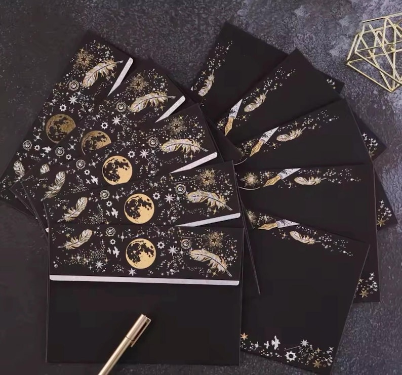 Puede incluir: Juego de papeler&iacute;a negro con dise&ntilde;os celestiales dorados. El conjunto incluye sobres y tarjetas adornados con lunas, plumas y estrellas. Un bol&iacute;grafo dorado descansa cerca de la papeler&iacute;a, sugiriendo un tema de escritura. La est&eacute;tica general es elegante y m&iacute;stica.