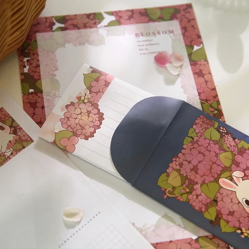 Pu&ograve; includere: Un set di cancelleria con un design floreale con ortensie rosa e un coniglio. La cancelleria include buste e carta da lettere. Il testo sulla carta recita "BLOSSOM".