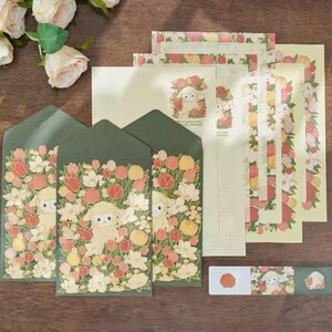 Pu&ograve; includere: Un set di cancelleria con un design floreale con un agnello al centro. Il set include buste, carta da lettere e un foglio di adesivi. Le buste sono verdi con un motivo floreale e un agnello al centro. La carta da lettere &egrave; crema con un bordo floreale e un agnello al centro. Il foglio di adesivi ha un design floreale con un agnello al centro.
