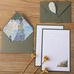 Pu&ograve; includere: Un set di cartoleria verde con un foglio di carta rigato e una busta con un dipinto di una donna con un ombrello. La busta ha una patta decorativa.