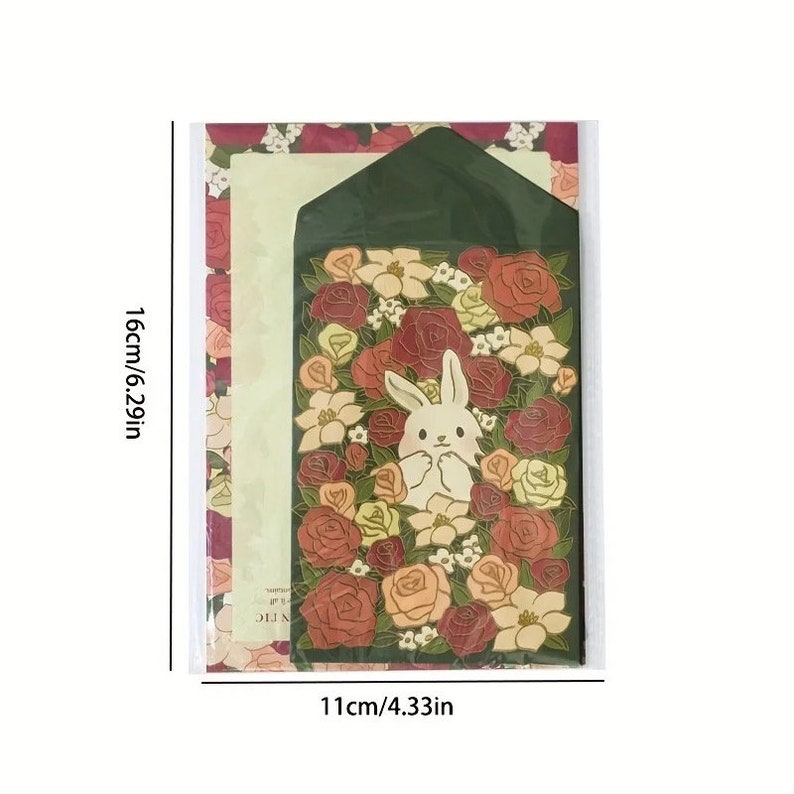Pu&ograve; includere: Un set di cancelleria con una busta verde con un coniglio bianco circondato da rose rosse, arancioni e gialle. La busta misura 11 cm di larghezza e 16 cm di altezza.