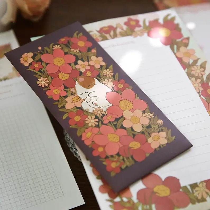 Pu&ograve; includere: Una busta viola con un disegno floreale che presenta un gatto bianco che dorme al centro. I fiori sono rosa, arancioni e gialli.
