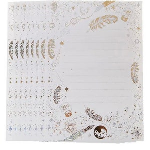 Puede incluir: Una pila de hojas de papeler&iacute;a blancas con un dise&ntilde;o celestial. Las hojas presentan detalles dorados y plateados de plumas, estrellas, lunas y tinteros. P&aacute;ginas rayadas listas para escribir.