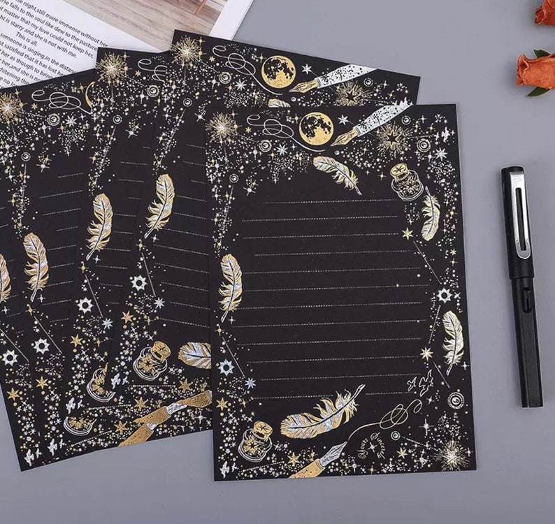 Puede incluir: Papel de papeler&iacute;a negro con detalles dorados. El dise&ntilde;o incluye plumas, estrellas, lunas, tinteros y plumas. Se ven p&aacute;ginas rayadas. Un bol&iacute;grafo negro est&aacute; en el lado derecho de la imagen.