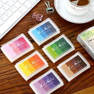 Könnte beinhalten: Sechs farbige Stempelkissen mit dem Text "Let's Color" auf jedem Kissen. Die Stempelkissen sind in einer Reihe auf einer Holzoberfläche angeordnet.