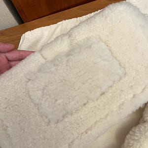 May include: Close-up of a cream-colored, fluffy, rectangular pillow or cushion. The surface has a textured, plush appearance, with a rectangular patch sewn onto the front. The item appears soft and comfortable.