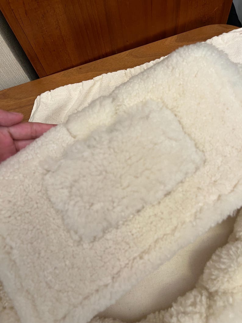 May include: Close-up of a cream-colored, fluffy, rectangular pillow or cushion. The surface has a textured, plush appearance, with a rectangular patch sewn onto the front. The item appears soft and comfortable.