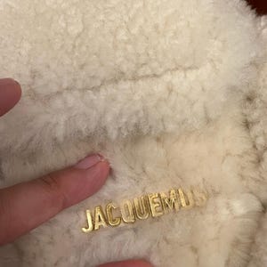 May include: Close-up of a cream-colored, textured fabric with the gold-colored word "JACQUEMUS" embossed on the surface. The fabric appears soft and fluffy, possibly wool or a similar material.