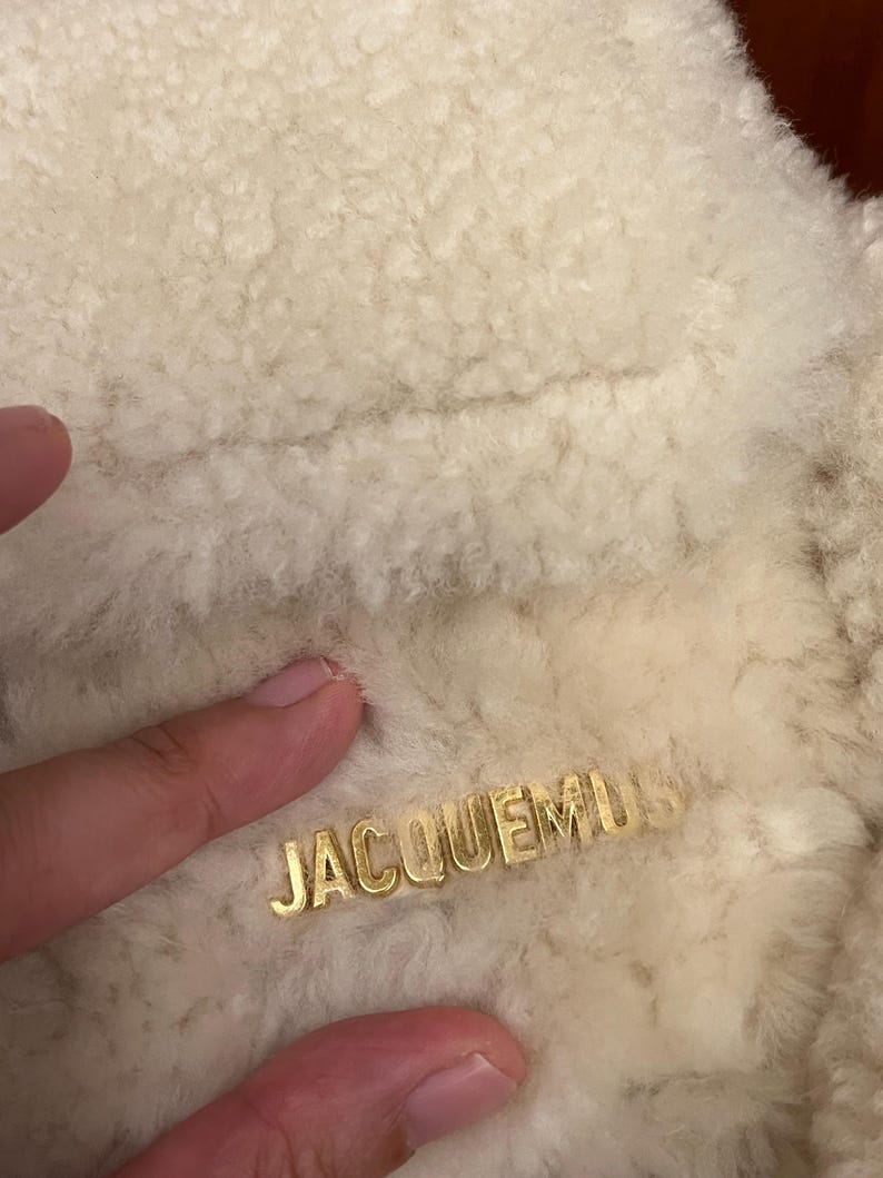 May include: Close-up of a cream-colored, textured fabric with the gold-colored word "JACQUEMUS" embossed on the surface. The fabric appears soft and fluffy, possibly wool or a similar material.