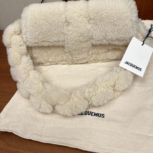 May include: Cream-colored, fluffy handbag with a braided handle. The bag has a rectangular shape with a flap closure. A tag with the brand name "JACQUEMUS" is attached. The bag rests on a matching fabric bag with the brand name printed on it.
