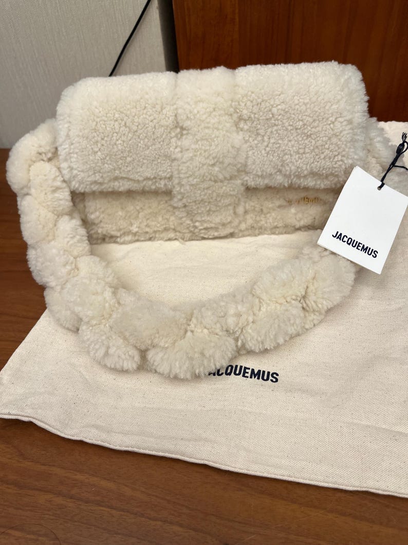 May include: Cream-colored, fluffy handbag with a braided handle. The bag has a rectangular shape with a flap closure. A tag with the brand name "JACQUEMUS" is attached. The bag rests on a matching fabric bag with the brand name printed on it.