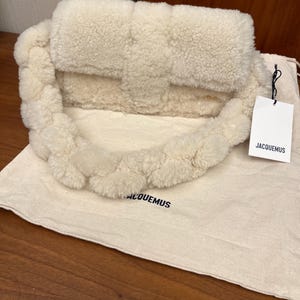 May include: Cream-colored, fluffy handbag with a braided strap. The bag has a rectangular shape with a flap closure. A tag with the brand name "JACQUEMUS" hangs from the bag. The bag rests on a matching fabric bag with the brand name printed on it.
