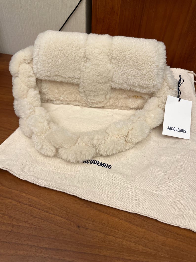 May include: Cream-colored, fluffy handbag with a braided strap. The bag has a rectangular shape with a flap closure. A tag with the brand name "JACQUEMUS" hangs from the bag. The bag rests on a matching fabric bag with the brand name printed on it.