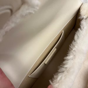 May include: Open view of a cream-colored handbag interior. The bag features a light-colored leather exterior and a fluffy, white, shearling-like lining. A small pocket is visible inside the bag. The bag is open, revealing the interior.