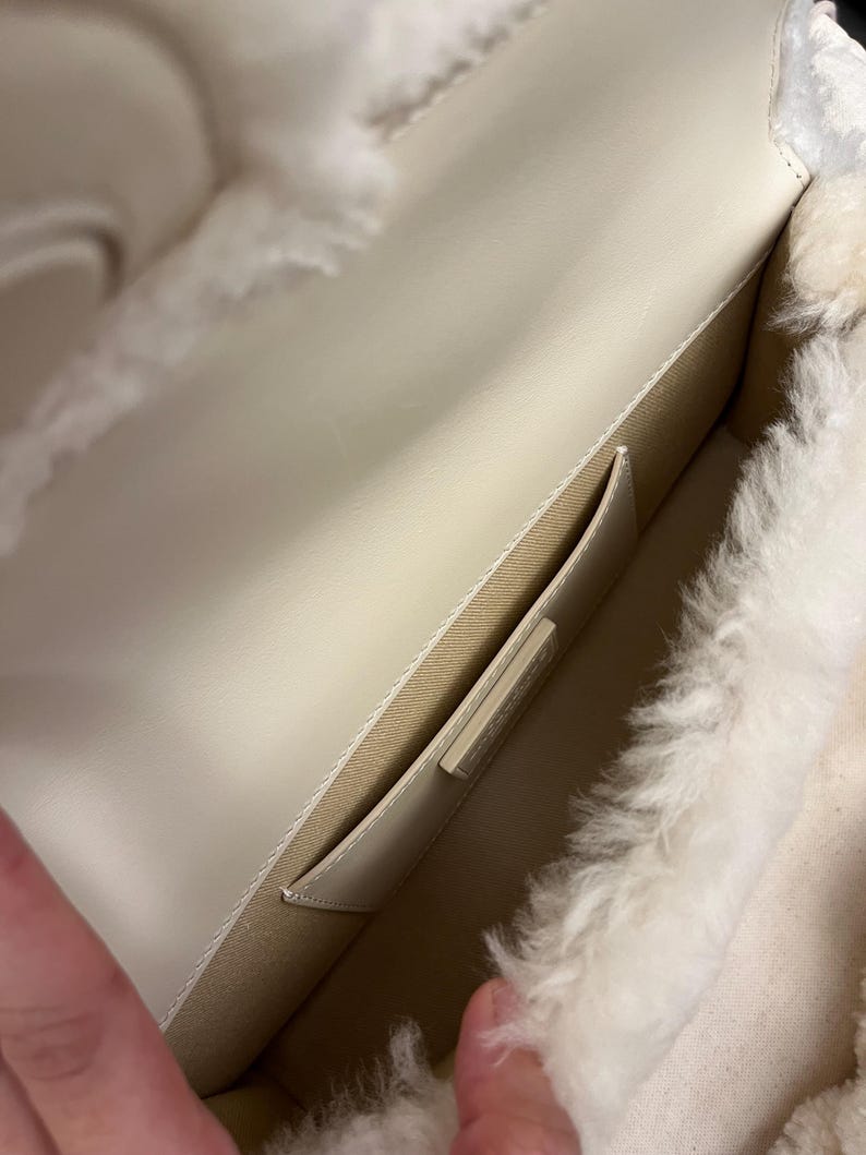 May include: Open view of a cream-colored handbag interior. The bag features a light-colored leather exterior and a fluffy, white, shearling-like lining. A small pocket is visible inside the bag. The bag is open, revealing the interior.