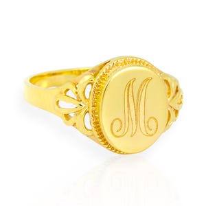 May include: A gold signet ring with the letter "M" engraved on a round face. The ring has a decorative floral design on the sides and a polished finish. The ring is a classic piece of jewelry.