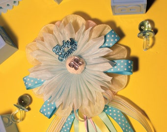 May include: A blue and white baby shower flower with a blue ribbon and white polka dots. The flower has a blue and silver "Baby Shower" charm in the centre. The flower is surrounded by yellow, blue and pink ribbons.