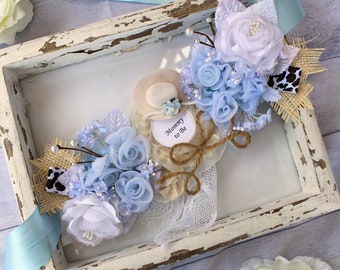May include: A white frame with a blue and white floral arrangement inside. The flowers are made of fabric and are arranged around a small, white hat with the words "Mommy to Be" written on it.