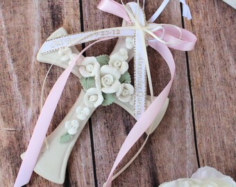 May include: A white ceramic cross with a floral design and a pink and white ribbon with the text "Alexander Garcia" and "Jn 14:6 9:23-12" on it.