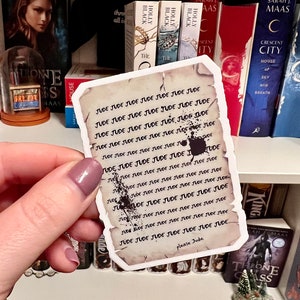 May include: A close-up of a sticker with the name "Jude" repeated many times in black ink on a parchment-like background. The sticker is being held by a hand with a light purple nail polish.