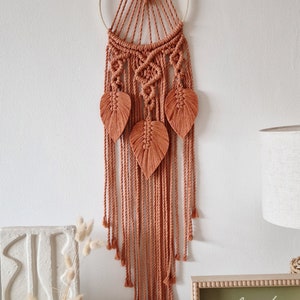 May include: A terracotta-coloured macrame wall hanging with a circular wooden hoop and three leaf-shaped accents. The hanging is decorated with a geometric pattern and has a "hello spring" sign below it.