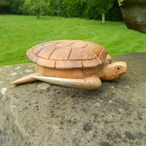 May include: A wooden turtle sculpture with a detailed shell and realistic features. The turtle is lying on a rock with a green grassy background.