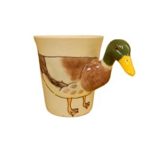 May include: A ceramic mug featuring a duck design. The beige mug has a brown and white duck illustration. The handle is a 3D duck head, with a green head and yellow beak. A unique kitchenware item.