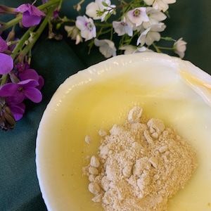 May include: A white bowl filled with a yellow liquid and a pile of beige powder. The bowl is on a green surface.