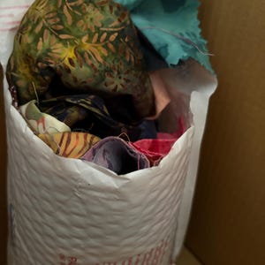 May include: A white padded envelope filled with fabric scraps. The fabrics include green, blue, and red pieces with floral and abstract patterns. The envelope is inside a cardboard box.