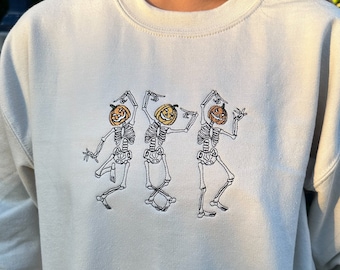 May include: A cream-colored sweatshirt with a graphic of three skeletons wearing pumpkin heads. The skeletons are dancing and have a fun, festive look.