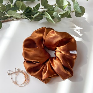 May include: A large, shiny, copper-colored satin scrunchie with a small white tag that reads "Koretty". A pair of hoop earrings with small pink beads are next to the scrunchie. Green leafy branches are in the background.