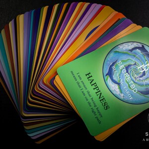 May include: A deck of colorful tarot cards with a green card on top that reads "SECRETBD A BLESSING TAROT" and "HAPPINESS I am aware that being happy means that I am on the right path".