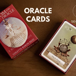 以下が含まれることがあります： 「Wisdom of the Oracle」と「A Blessing Tarot and Guidance Deck」by Colette Baron-Reidと書かれた赤い箱。箱が開いており、時計と熱気球が描かれたカードが見えています。カードには「Tick-Tock」という文字が巻物に書かれています。