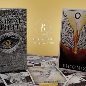 May include: A box of Animal Spirit Tarot cards by Kim Krans, featuring a silver and black design with a large eye on the front. The box is sitting on a table with a spread of tarot cards, including the Phoenix card, which features a white figure with wings against a red and orange background.