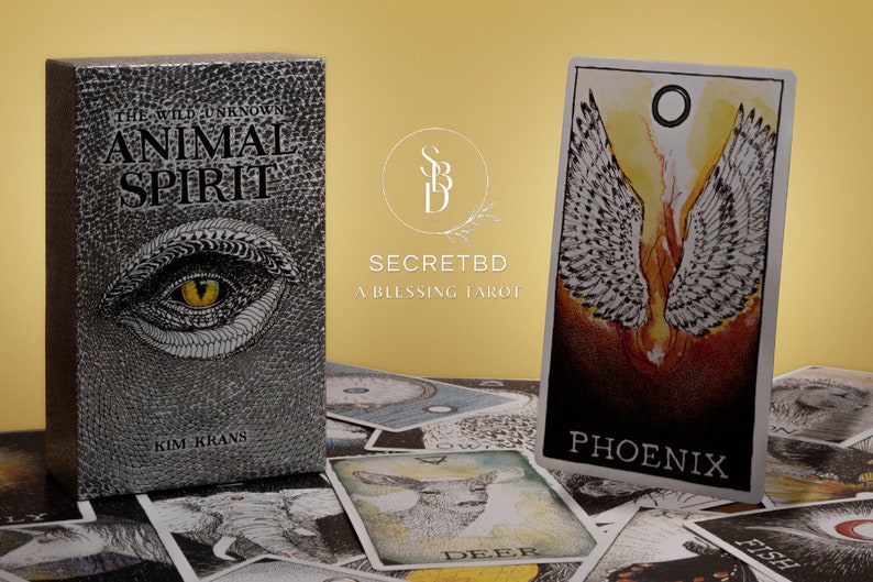 May include: A box of Animal Spirit Tarot cards by Kim Krans, featuring a silver and black design with a large eye on the front. The box is sitting on a table with a spread of tarot cards, including the Phoenix card, which features a white figure with wings against a red and orange background.