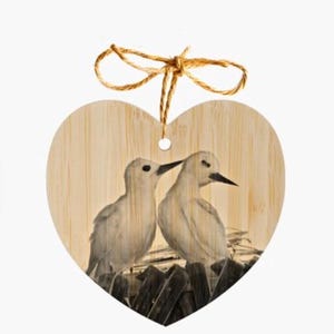 May include: A wooden heart-shaped ornament featuring a painted image of two white birds perched on a nest. The birds are facing each other and appear to be interacting.